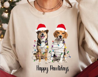 May include: Beige sweatshirt with a festive design featuring two dogs in Santa hats, wrapped in Christmas lights. The words "Happy Pawlidays" are printed below. A seasonal, holiday-themed garment.