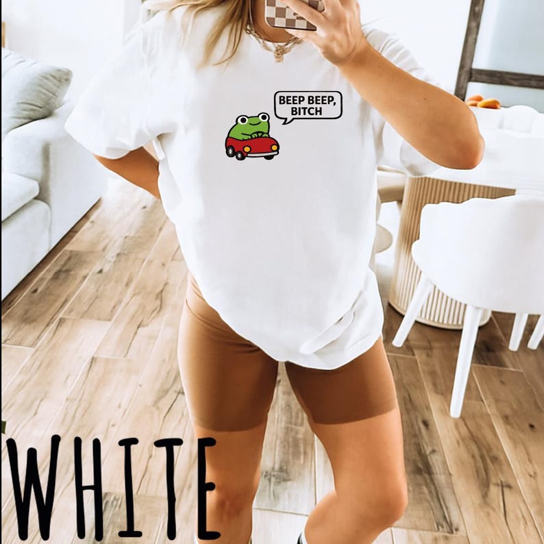 A person is taking a selfie in a room with a white t-shirt featuring a frog graphic and text.