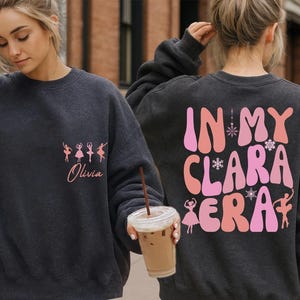 May include: Dark gray sweatshirt with a ballet theme. The front features a pink graphic of ballerinas and the name "Olivia." The back displays the text "IN MY CLARA ERA" in pink and orange, with snowflake and ballerina graphics.