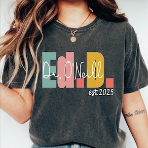 May include: A dark gray t-shirt with a colorful graphic design that reads "Dr. O'Neill est. 2025". The graphic is made up of colorful blocks with white lettering.