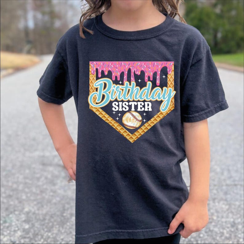 A person wearing a black t-shirt with a graphic design that says "Birthday Sister" and features a baseball and a waffle cone.