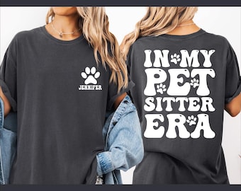 May include: A dark gray t-shirt with a white paw print and the name "Jennifer" on the front. The back of the shirt has a white graphic that says "In My Pet Sitter Era" with paw prints.
