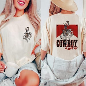 May include: Two women wearing cream colored t-shirts with a skeleton cowboy riding a horse graphic. The shirt on the right has the text "COWBOY RIDERS" in red and black.