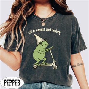 May include: Dark gray t-shirt with the text "Off to commit aunt foolery." The shirt features a green frog wearing a wizard hat and riding a scooter. The frog graphic is in a whimsical style.