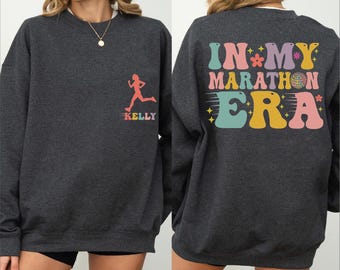 May include: A dark gray crewneck sweatshirt featuring a small pink runner silhouette and the name Kelly on the front left chest. The back displays the colorful text IN MY MARATHON ERA with a disco ball and star accents in pastel tones.