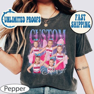 Pu&ograve; includere: Una maglietta nera con un design personalizzato che presenta un gruppo di ragazze in uniformi da cheerleader con pompon. Il design include la parola "CUSTOM" in un gradiente di colori e il nome "Olivia" in un font scritto a mano.