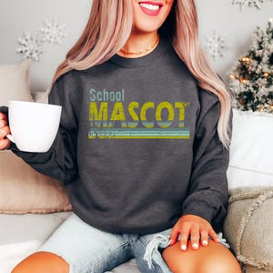 May include: Dark gray sweatshirt with the text "School MASCOT" in distressed yellow and teal lettering. The sweatshirt has a crew neck and long sleeves. The design includes horizontal stripes in teal and yellow.