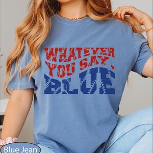 May include: A light blue t-shirt with the text "WHATEVER YOU SAY, BLUE" in red and blue. The word "BLUE" has a baseball stitch design. The shirt is a casual style.