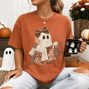 May include: A rust-colored short-sleeve t-shirt featuring a central graphic of a ghost wearing a leopard print bow and holding a pumpkin. The ghost is decorated with various patchwork patterns including plaid and floral designs. The shirt is paired with light blue distressed denim jeans.
