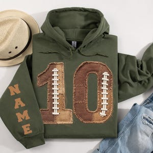 May include: An olive green hooded sweatshirt featuring a large number 10 on the front, designed with brown textured patches and white lacing to resemble a football. The word NAME is printed in brown on the sleeve, accompanied by a straw fedora hat.