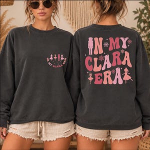 May include: A charcoal gray crewneck sweatshirt featuring a small graphic on the front left chest with the text "IN MY CLARA ERA" and ballet figures. The back displays a large graphic with the same text, a nutcracker, and ballet dancers.