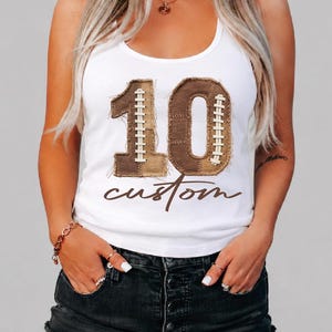 May include: A white tank top featuring a large graphic of the number 10 in a brown, textured football-inspired pattern with white lace-up details. Below the number, the word custom is printed in a brown, cursive script font across the chest.