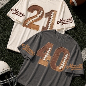 May include: Two short-sleeve crew neck t-shirts, one cream and one charcoal gray. Each features large, frayed-edge brown fabric numbers on the front, stitched with white laces. The word Name is on the left sleeve and Mascot is on the right.