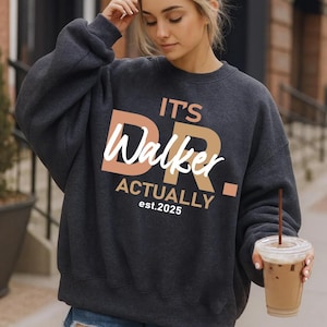 May include: Dark gray sweatshirt with the text "It's Dr. Walker Actually est. 2025" in white and peach lettering. The sweatshirt is a casual style, perfect for everyday wear.