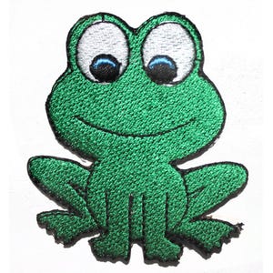 May include: Embroidered patch of a smiling green frog with large white eyes and blue pupils. The frog has a black outline and is sitting upright, with its legs and feet visible. The patch is likely for clothing or accessories.