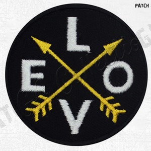 Op de afbeelding: Zwarte ronde patch met het woord "LOVE" in wit geborduurd, doorsneden door twee gele pijlen. De patch is een decoratief item, waarschijnlijk voor kleding of accessoires.