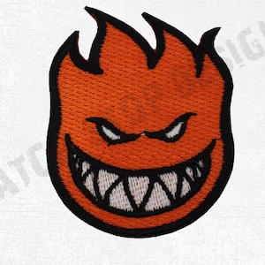 Könnte beinhalten: Gestickter Aufnäher mit einer stilisierten orangefarbenen Flamme mit grinsendem Gesicht, schwarzer Umrandung und weißen Zähnen. Der Aufnäher hat ein strukturiertes Aussehen und ist wahrscheinlich für das Aufnähen auf Kleidung oder Accessoires gedacht.