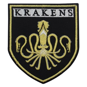 Könnte beinhalten: Schwarzer Aufnäher mit einem goldgestickten Kraken-Design. Das Wort "KRAKENS" ist in Weiß auf einem rechteckigen Banner gestickt. Der Aufnäher hat einen goldenen Rand und eine Schildform.