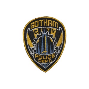 Może przedstawiać: Haftowana naszywka Gotham City Police Dept. Naszywka ma kształt tarczy z czarnym tłem i złotą obwódką. Tekst "Gotham City Police Dept." jest wyświetlany złotymi literami. Środek przedstawia stylizowany krajobraz miejski w kolorze niebieskim i złotym.