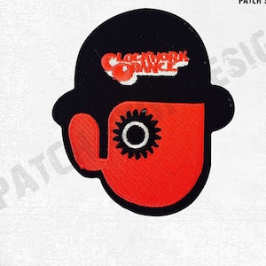 Puede incluir: Parche bordado con un sombrero negro y una cara roja con un engranaje. El texto "CLOCKWORK ORANGE" está bordado en blanco y naranja. Este parche está diseñado para ropa o accesorios.