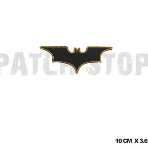 Puede incluir: Parche negro con forma de murciélago y ribete dorado, que mide aproximadamente 10 cm de ancho y 3,6 cm de alto. El parche tiene un diseño bordado detallado. El texto "PATCH STOP" es visible en el fondo.