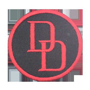 May include: A round, black patch with a red border. The patch features the intertwined red letters "DD" in a bold, stylized font. The design is embroidered on the fabric.