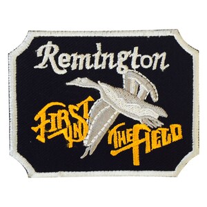 May include: Embroidered patch with a black background and a white border. The patch features the word "Remington" in white script above a flying bird. Below the bird, the words "FIRST IN THE FIELD" are in yellow.