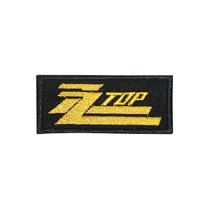 May include: A rectangular black patch with the words "ZZ TOP" embroidered in gold thread. The design features a stylized "ZZ" and the word "TOP" in a bold, sans-serif font. The patch is likely for clothing or accessories.