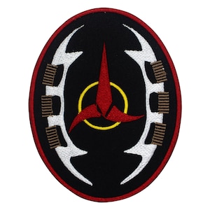 Puede incluir: Parche bordado de forma ovalada, con fondo negro y emblema rojo y amarillo. El emblema presenta una forma roja de tres puntas dentro de un círculo amarillo. Formas blancas y curvas con detalles marrones bordean el emblema, y el parche tiene un borde rojo.