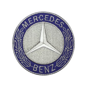 Könnte beinhalten: Runder, gestickter Aufnäher mit dem Mercedes-Benz-Logo. Der Aufnäher hat einen blauen Rand mit den weißen Schriftzügen "MERCEDES" und "BENZ". In der Mitte befindet sich ein silberner Stern in einem silbernen Kreis, umgeben von einem Lorbeerkranz.