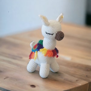 May include: White crocheted llama toy with a rainbow colored fringe around its neck. The llama has brown eyes and a brown nose.