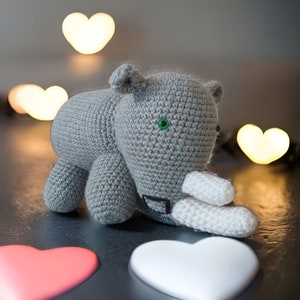 May include: A gray crocheted rhinoceros toy with a white crocheted horn. The toy has green eyes and is lying on a dark surface with white hearts in the background.