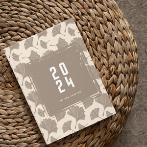 Puede incluir: Una agenda beige de 2024 con un patrón de hojas botánicas y el texto "2024 My New Chapter" en blanco y negrita. La agenda está sobre una superficie de mimbre tejido.