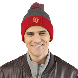 May include: A grey and red knitted beanie hat with a pom-pom. The red band has the embroidered number '67' in yellow and pink. This winter hat is a stylish accessory.