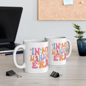 May include: Two white ceramic mugs with the text "IN MY MAHJONG ERA" in a retro font. The text is in shades of blue, pink, and orange with decorative elements. The mugs are on a light-colored surface.