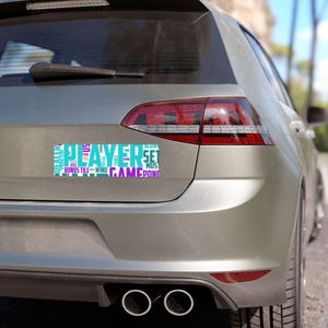 May include: A rectangular car bumper sticker with the word "PLAYER" in large, light blue letters. Other words like "BONUS", "TILE", "WIND", "GAME", and "SET" are in various colours and fonts. The sticker is affixed to a silver car.