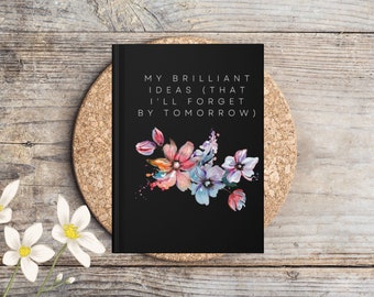 May include: Black journal with the text "MY BRILLIANT IDEAS (THAT I'LL FORGET BY TOMORROW)" and a watercolor floral design in pink, blue, and purple. The journal is on a cork coaster, with white flowers in the foreground.