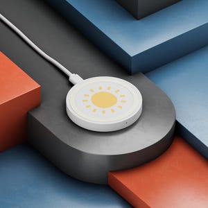 Op de afbeelding: Een witte draadloze oplader met een gele zon. De ronde oplader heeft een witte kabel. De achtergrond bevat geometrische vormen in rood, grijs en blauw.
