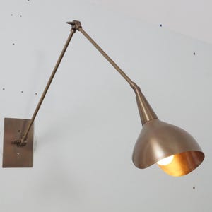 Può includere: Un apparecchio di illuminazione a parete in ottone con un braccio regolabile e un paralume a forma di campana. La luce è accesa e illumina la stanza.