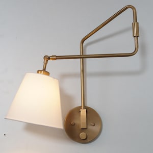 Puede incluir: Una lámpara de pared de latón con una pantalla de tela blanca. La lámpara tiene un diseño geométrico con un brazo largo que se extiende desde la pared y un brazo más corto que sostiene la pantalla. La lámpara tiene un interruptor en la placa de pared.