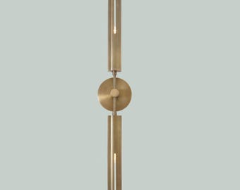 Puede incluir: Una lámpara de pared de latón con una sección central circular y dos secciones verticales con una ranura en el medio. La lámpara tiene un diseño minimalista.
