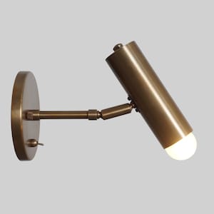 May include: A brass wall-mounted light fixture with a cylindrical shade and an adjustable arm. The light is turned on and a white light bulb is visible.