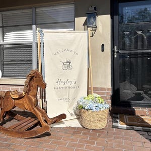 May include: A wooden rocking horse and a baby shower welcome sign with the text "Welcome to Hayley's Baby Shower" are displayed outside a home. A woven basket of blue and green flowers sits nearby.