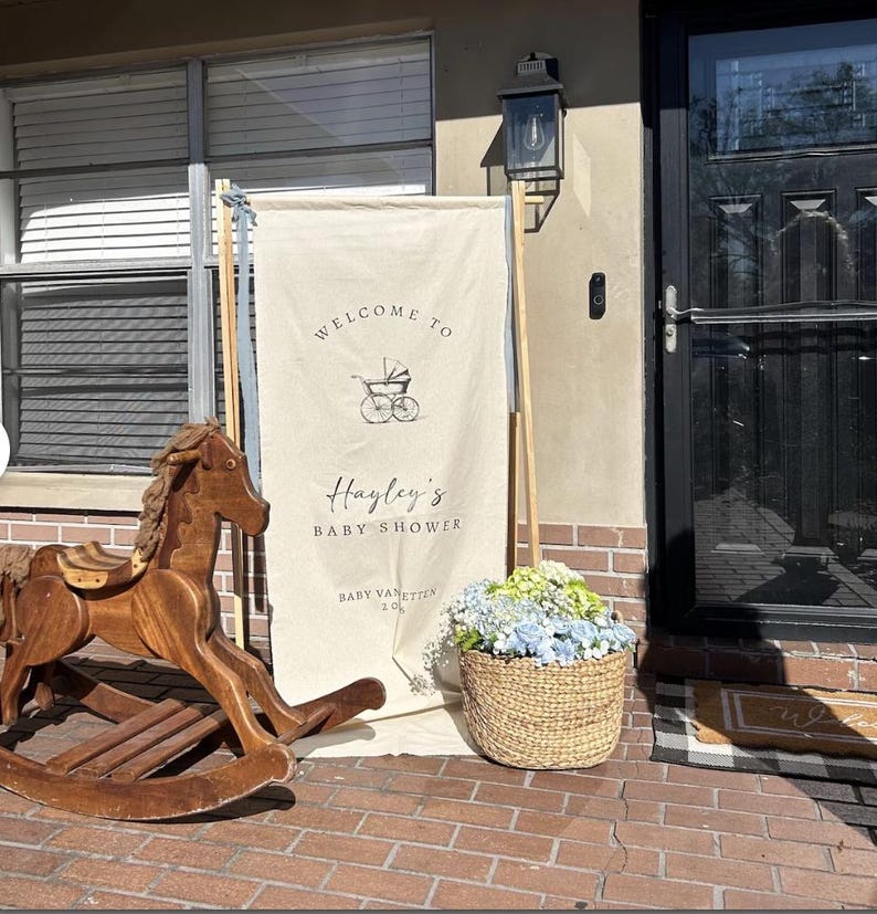 May include: A wooden rocking horse and a baby shower welcome sign with the text "Welcome to Hayley's Baby Shower" are displayed outside a home. A woven basket of blue and green flowers sits nearby.