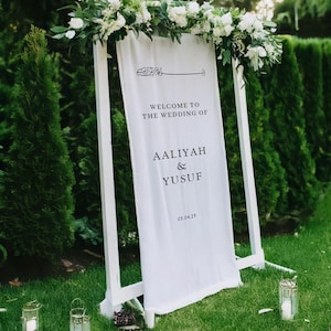 May include: White wedding sign with calligraphy and the text "Welcome to the wedding of Aaliyah & Yusuf 05.04.25" on a white background. The sign is decorated with white flowers and greenery.
