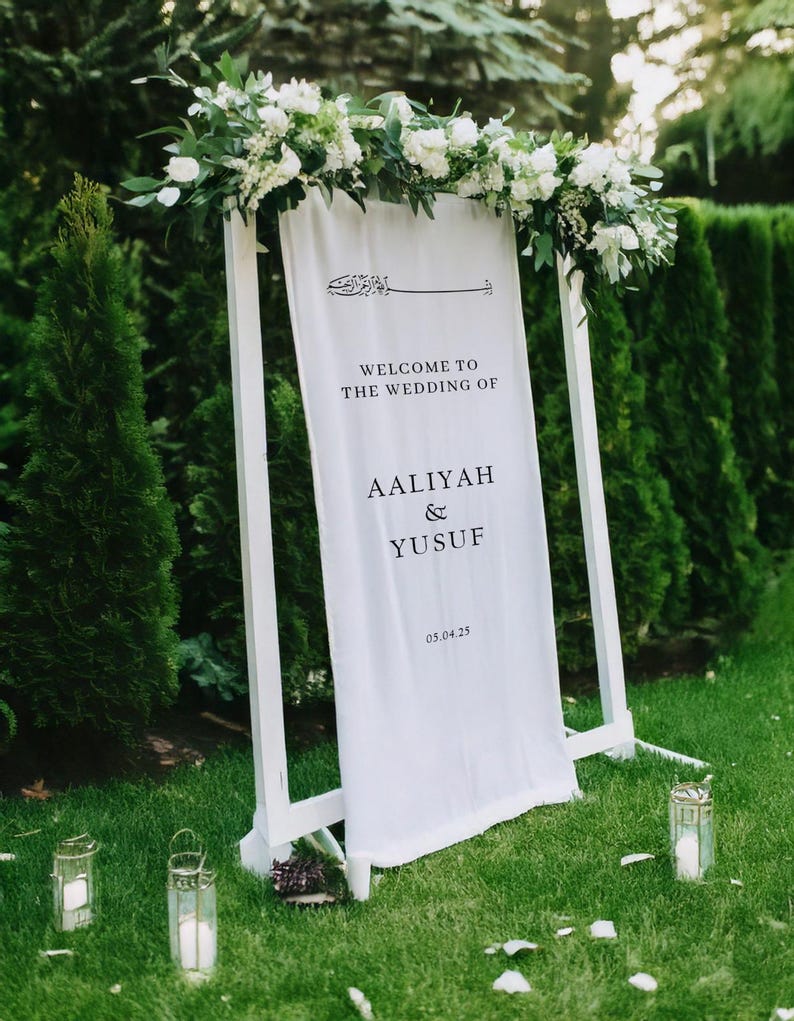May include: White wedding sign with calligraphy and the text "Welcome to the wedding of Aaliyah & Yusuf 05.04.25" on a white background. The sign is decorated with white flowers and greenery.