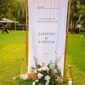 May include: White wedding sign with black text that reads "Welcome to the wedding of Jasmine & Kareem 29.03.25". The sign is standing on a wooden frame with greenery and white flowers in front of it.