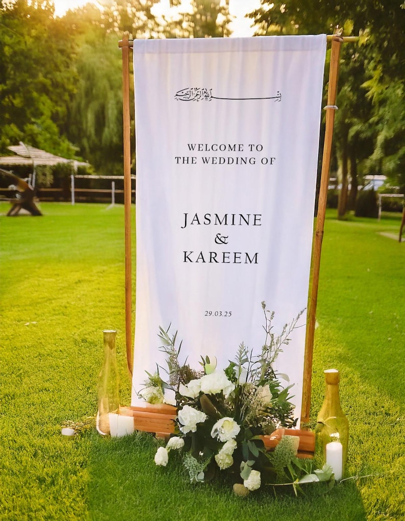 May include: White wedding sign with black text that reads "Welcome to the wedding of Jasmine & Kareem 29.03.25". The sign is standing on a wooden frame with greenery and white flowers in front of it.