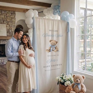 May include: A vertical fabric banner for a baby shower featuring a brown teddy bear illustration with a blue ribbon, the text WE CAN Bearly Wait WELCOME TO July's BABY SHOWER, and light blue fabric drapes accented with white and light blue balloons.