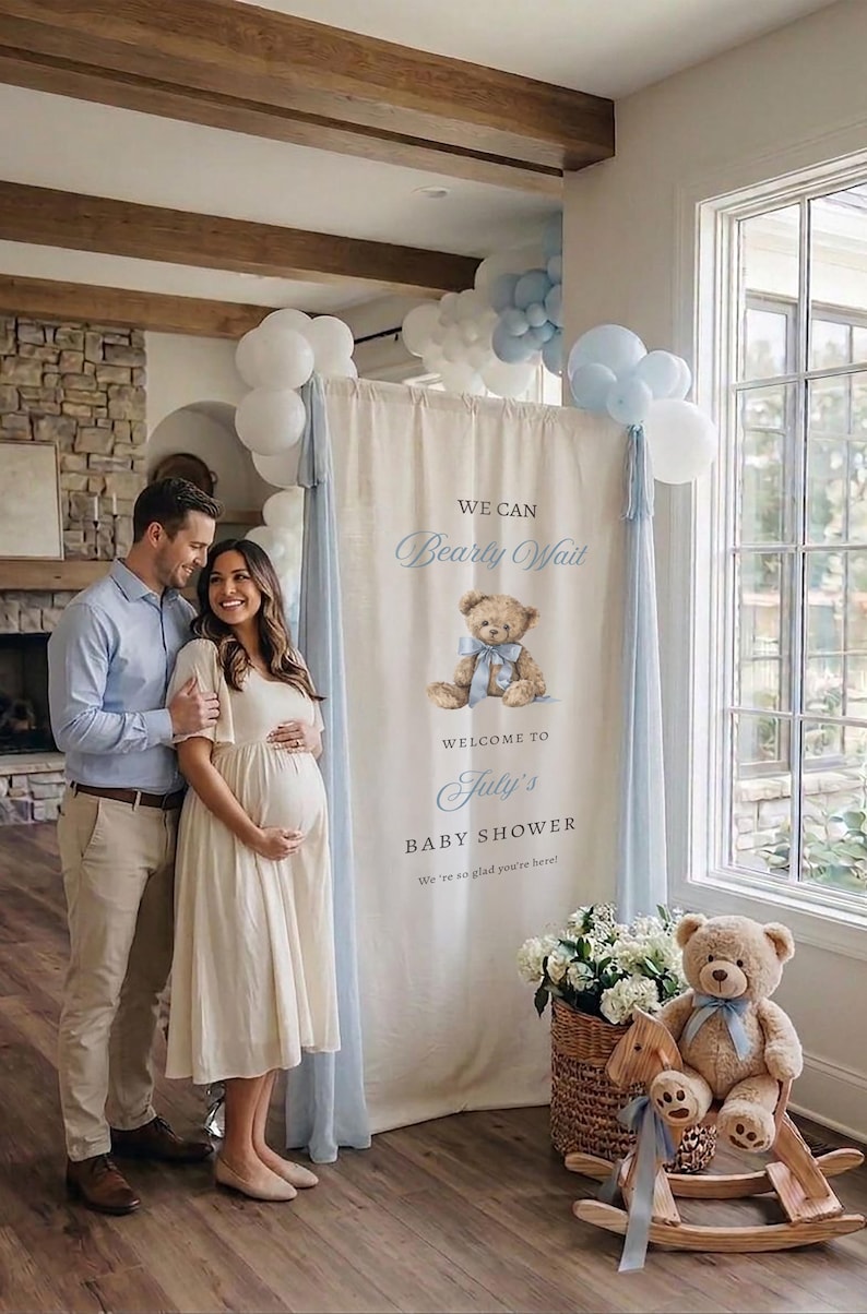 May include: A vertical fabric banner for a baby shower featuring a brown teddy bear illustration with a blue ribbon, the text WE CAN Bearly Wait WELCOME TO July's BABY SHOWER, and light blue fabric drapes accented with white and light blue balloons.